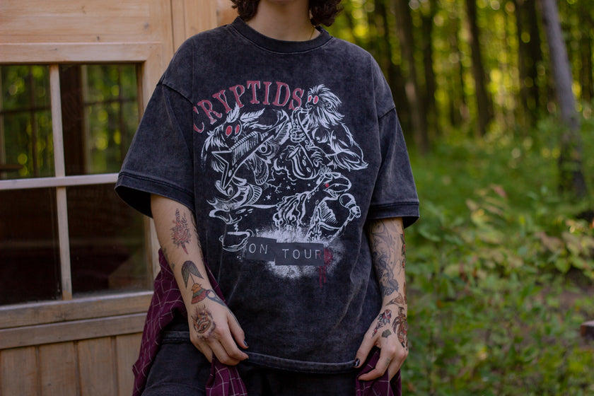 Black acid-washed oversized 80's style cotton band t-shirt with a vintage-style graphic depicting cryptids, including Bigfoot and Mothman, along with the bold text 'Cryptids On Tour' in red and white, and a list of shows across various locations and dates on the back.