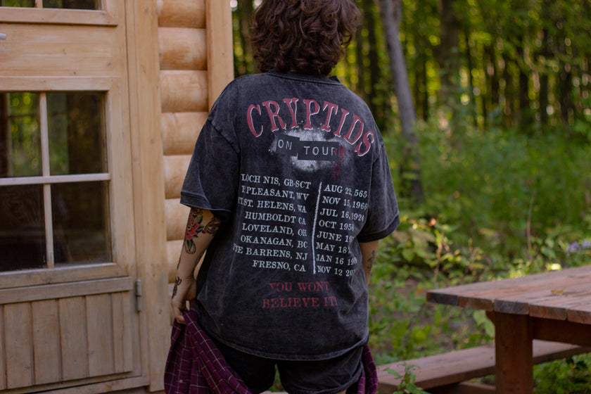 Black acid-washed oversized 80's style cotton band t-shirt with a vintage-style graphic depicting cryptids, including Bigfoot and Mothman, along with the bold text 'Cryptids On Tour' in red and white, and a list of shows across various locations and dates on the back.