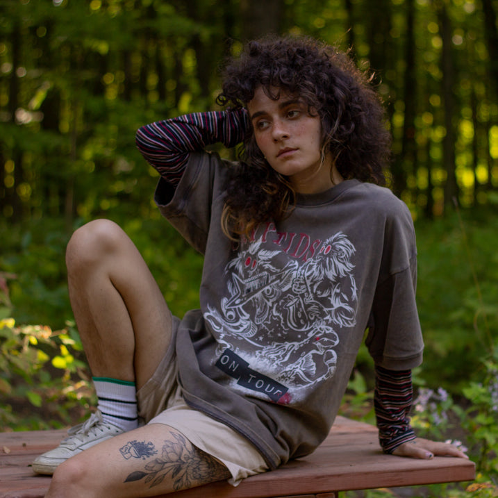 Bone-dust brown oversized cotton 80â€™s style band t-shirt with a vintage-style graphic depicting cryptids, including Bigfoot and Mothman, along with the bold text 'Cryptids On Tour' in red and white, and a list of shows across various locations and dates on the back.