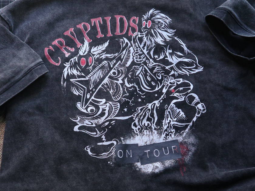 Black acid-washed oversized 80's style cotton band t-shirt with a vintage-style graphic depicting cryptids, including Bigfoot and Mothman, along with the bold text 'Cryptids On Tour' in red and white, and a list of shows across various locations and dates on the back.