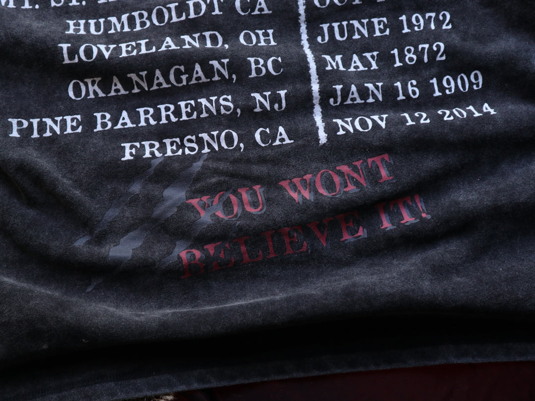 Black acid-washed oversized 80's style cotton band t-shirt with a vintage-style graphic depicting cryptids, including Bigfoot and Mothman, along with the bold text 'Cryptids On Tour' in red and white, and a list of shows across various locations and dates on the back.