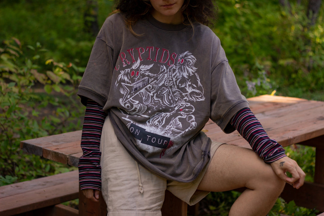 Bone-dust brown oversized cotton 80â€™s style band t-shirt with a vintage-style graphic depicting cryptids, including Bigfoot and Mothman, along with the bold text 'Cryptids On Tour' in red and white, and a list of shows across various locations and dates on the back.