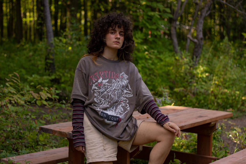 Bone-dust brown oversized cotton 80â€™s style band t-shirt with a vintage-style graphic depicting cryptids, including Bigfoot and Mothman, along with the bold text 'Cryptids On Tour' in red and white, and a list of shows across various locations and dates on the back.