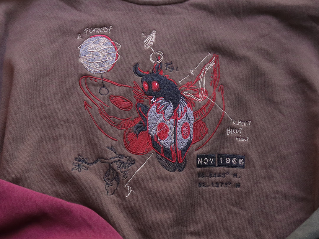 Cozy brown, green, and burgundy cotton crewneck sweatshirt, featuring a vintage, 80â€™s summer camp style embroidered design of the Mothman with cryptid-themed symbols and text.