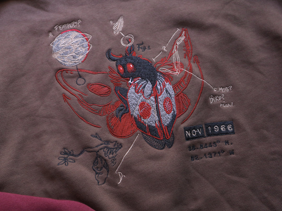 Cozy brown, green, and burgundy cotton crewneck sweatshirt, featuring a vintage, 80â€™s summer camp style embroidered design of the Mothman with cryptid-themed symbols and text.