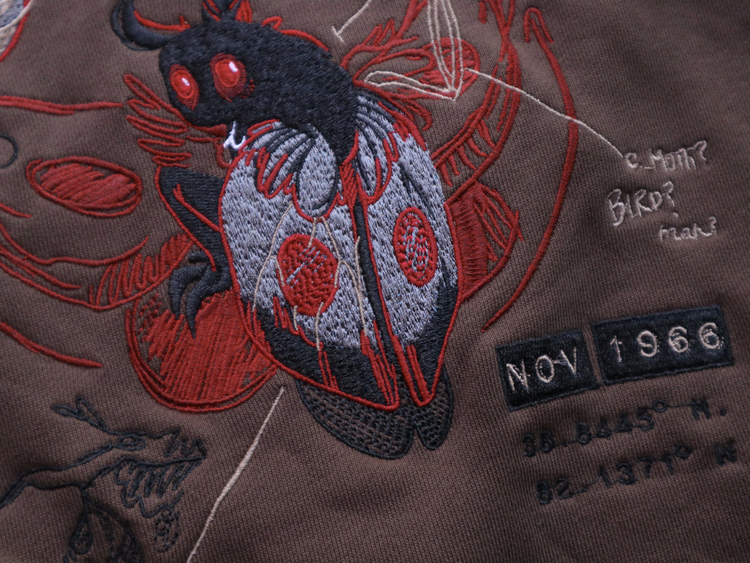 Cozy brown, green, and burgundy cotton crewneck sweatshirt, featuring a vintage, 80â€™s summer camp style embroidered design of the Mothman with cryptid-themed symbols and text.
