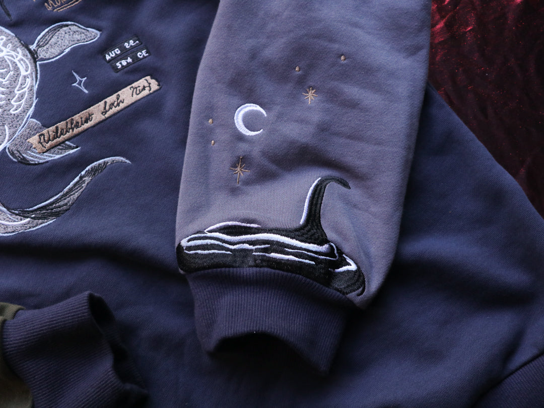 Cozy navy and fog-blue cotton crewneck sweatshirt, featuring a vintage, 80â€™s summer camp style embroidered design of the Mothman with cryptid-themed symbols and text.