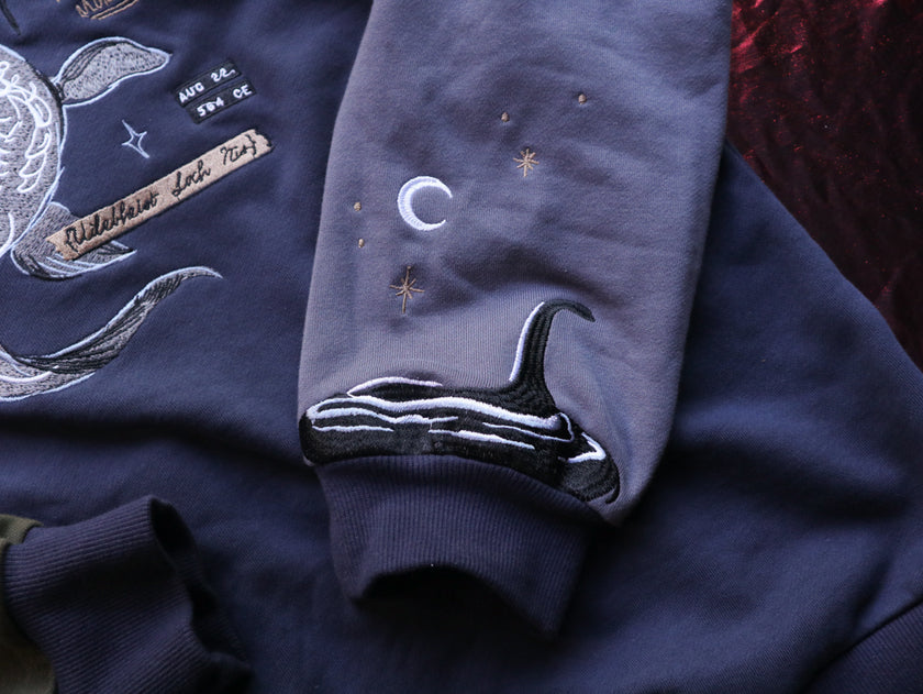 Cozy navy and fog-blue cotton crewneck sweatshirt, featuring a vintage, 80â€™s summer camp style embroidered design of the Mothman with cryptid-themed symbols and text.