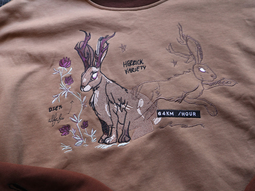 Cozy rust-orange, and peach cotton crewneck sweatshirt, featuring a vintage, embroidered design of the Jackalope with cryptid summer camp-themed symbols and text.