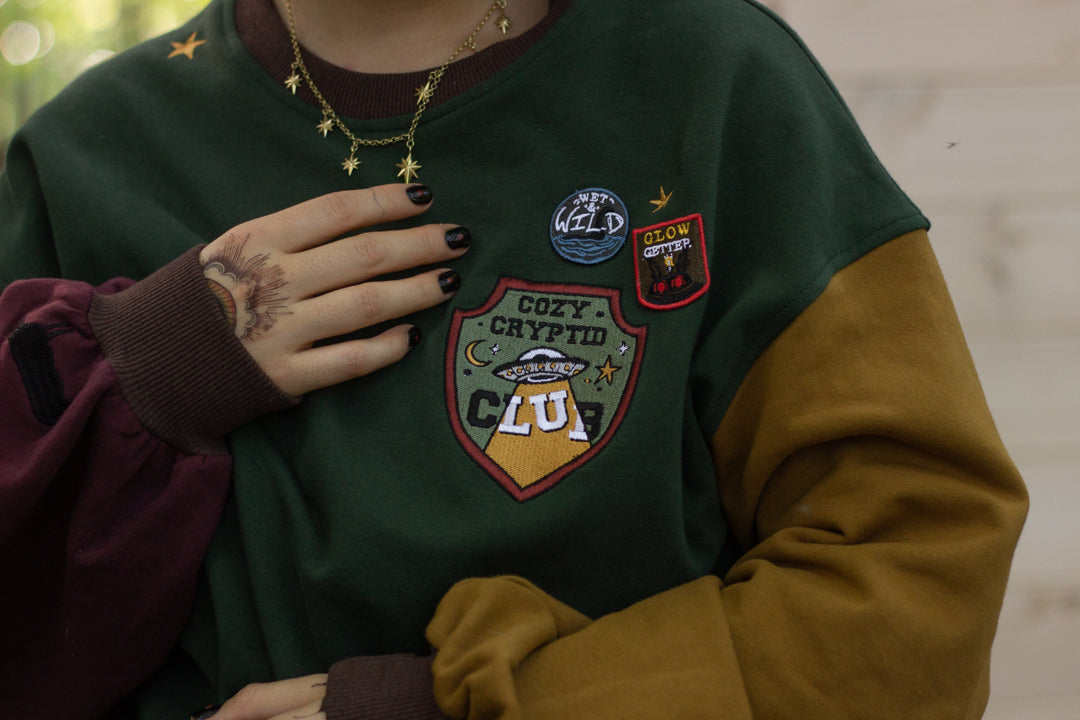 Cozy Forest green, deep yellow, and burgundy cotton crewneck sweatshirt, featuring a vintage, 80â€™s summer camp style embroidered â€œiron onâ€ patches, designed with cryptid-themed symbols and text.