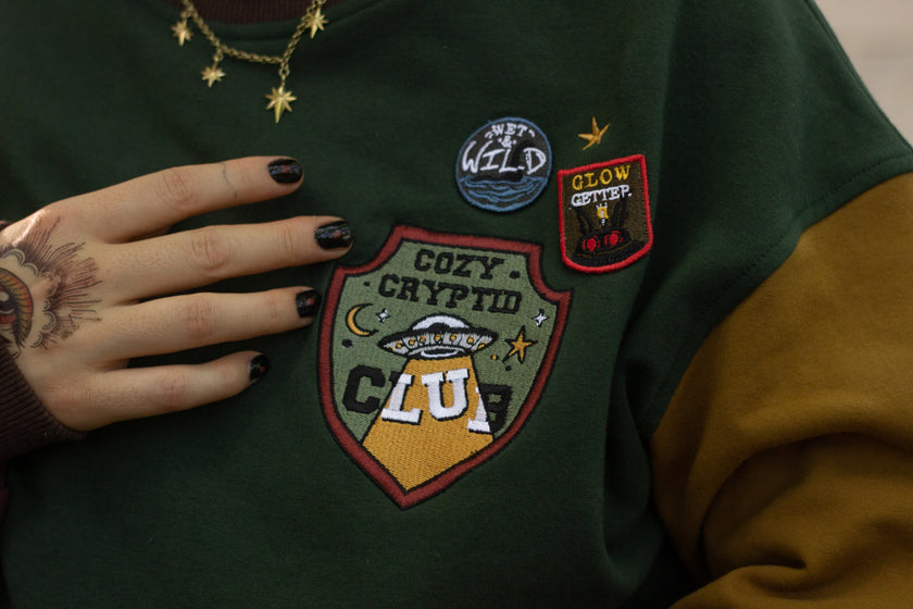Cozy Forest green, deep yellow, and burgundy cotton crewneck sweatshirt, featuring a vintage, 80â€™s summer camp style embroidered â€œiron onâ€ patches, designed with cryptid-themed symbols and text.