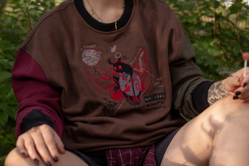 Cozy brown, green, and burgundy cotton crewneck sweatshirt, featuring a vintage, 80â€™s summer camp style embroidered design of the Mothman with cryptid-themed symbols and text.