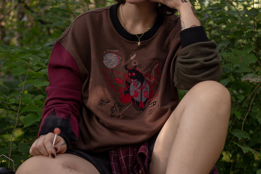 Cozy brown, green, and burgundy cotton crewneck sweatshirt, featuring a vintage, 80â€™s summer camp style embroidered design of the Mothman with cryptid-themed symbols and text.