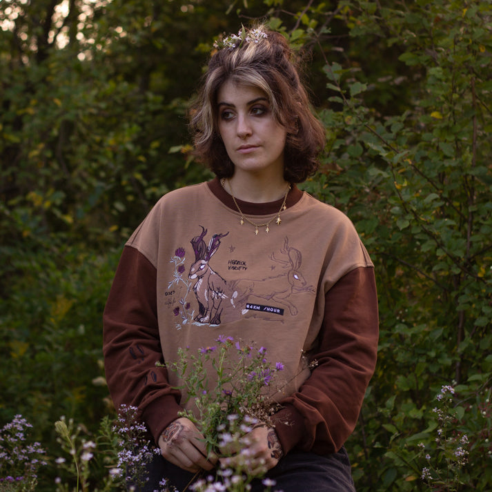 Cozy rust-orange, and peach cotton crewneck sweatshirt, featuring a vintage, embroidered design of the Jackalope with cryptid summer camp-themed symbols and text.