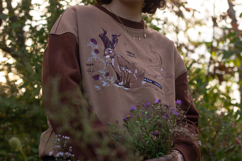 Cozy rust-orange, and peach cotton crewneck sweatshirt, featuring a vintage, embroidered design of the Jackalope with cryptid summer camp-themed symbols and text.