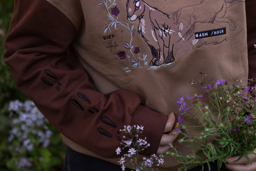 Cozy rust-orange, and peach cotton crewneck sweatshirt, featuring a vintage, embroidered design of the Jackalope with cryptid summer camp-themed symbols and text.