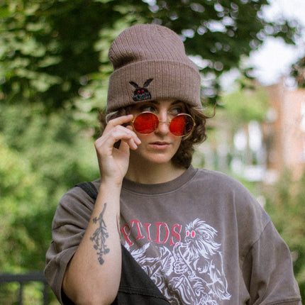 A cozy bone-dust brown cryptid-themed knit wool beanie with colourful speckles, embroidered with a tiny red-eyed, black-winged, flame-loving moth, and a secret message.