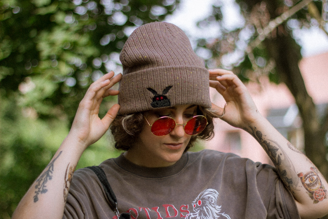 A cozy bone-dust brown cryptid-themed knit wool beanie with colourful speckles, embroidered with a tiny red-eyed, black-winged, flame-loving moth, and a secret message.