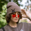 A cozy bone-dust brown cryptid-themed knit wool beanie with colourful speckles, embroidered with a tiny red-eyed, black-winged, flame-loving moth, and a secret message.