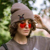 A cozy bone-dust brown cryptid-themed knit wool beanie with colourful speckles, embroidered with a tiny red-eyed, black-winged, flame-loving moth, and a secret message.