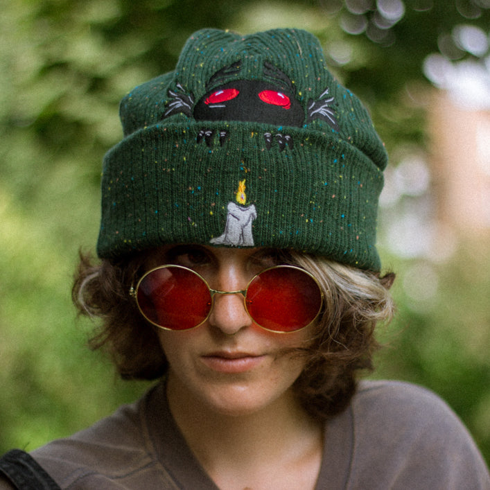 A cozy goblin-green knit wool beanie with colourful speckles, embroidered with a red-eyed, black-winged, flame-loving moth, and a glowing candle.