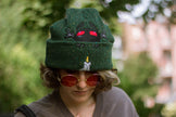 A cozy goblin-green knit wool beanie with colourful speckles, embroidered with a red-eyed, black-winged, flame-loving moth, and a glowing candle.