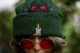 A cozy goblin-green knit wool beanie with colourful speckles, embroidered with a red-eyed, black-winged, flame-loving moth, and a glowing candle.