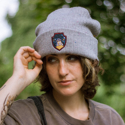 A cozy fog-grey cryptid camp-themed knit wool beanie with colourful speckles, embroidered with a cryptid club “iron on” patch, and Nessie monster.