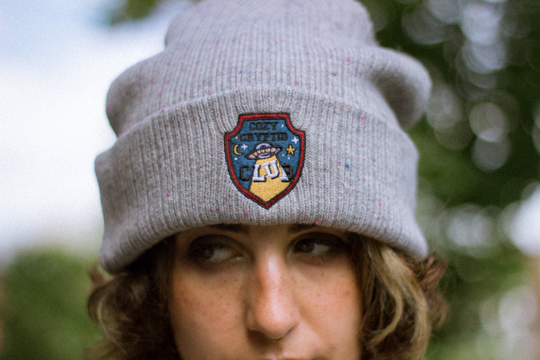 A cozy fog-grey cryptid camp-themed knit wool beanie with colourful speckles, embroidered with a cryptid club “iron on” patch, and Nessie monster.