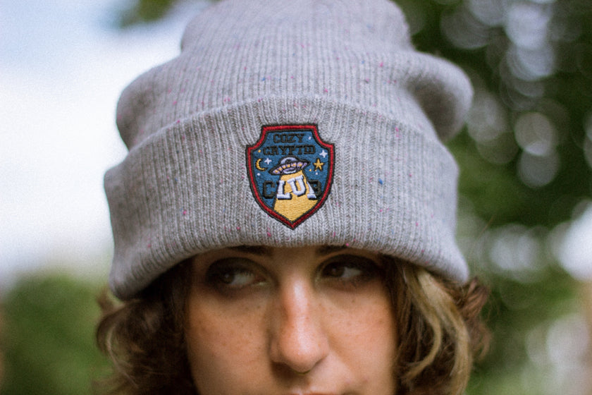 A cozy fog-grey cryptid camp-themed knit wool beanie with colourful speckles, embroidered with a cryptid club “iron on” patch, and Nessie monster.