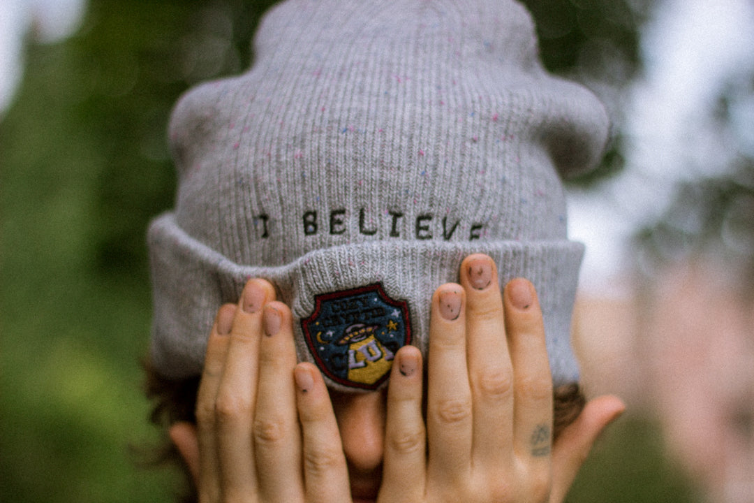 A cozy fog-grey cryptid camp-themed knit wool beanie with colourful speckles, embroidered with a cryptid club “iron on” patch, and Nessie monster.