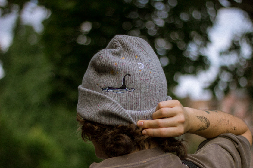 A cozy fog-grey cryptid camp-themed knit wool beanie with colourful speckles, embroidered with a cryptid club “iron on” patch, and Nessie monster.