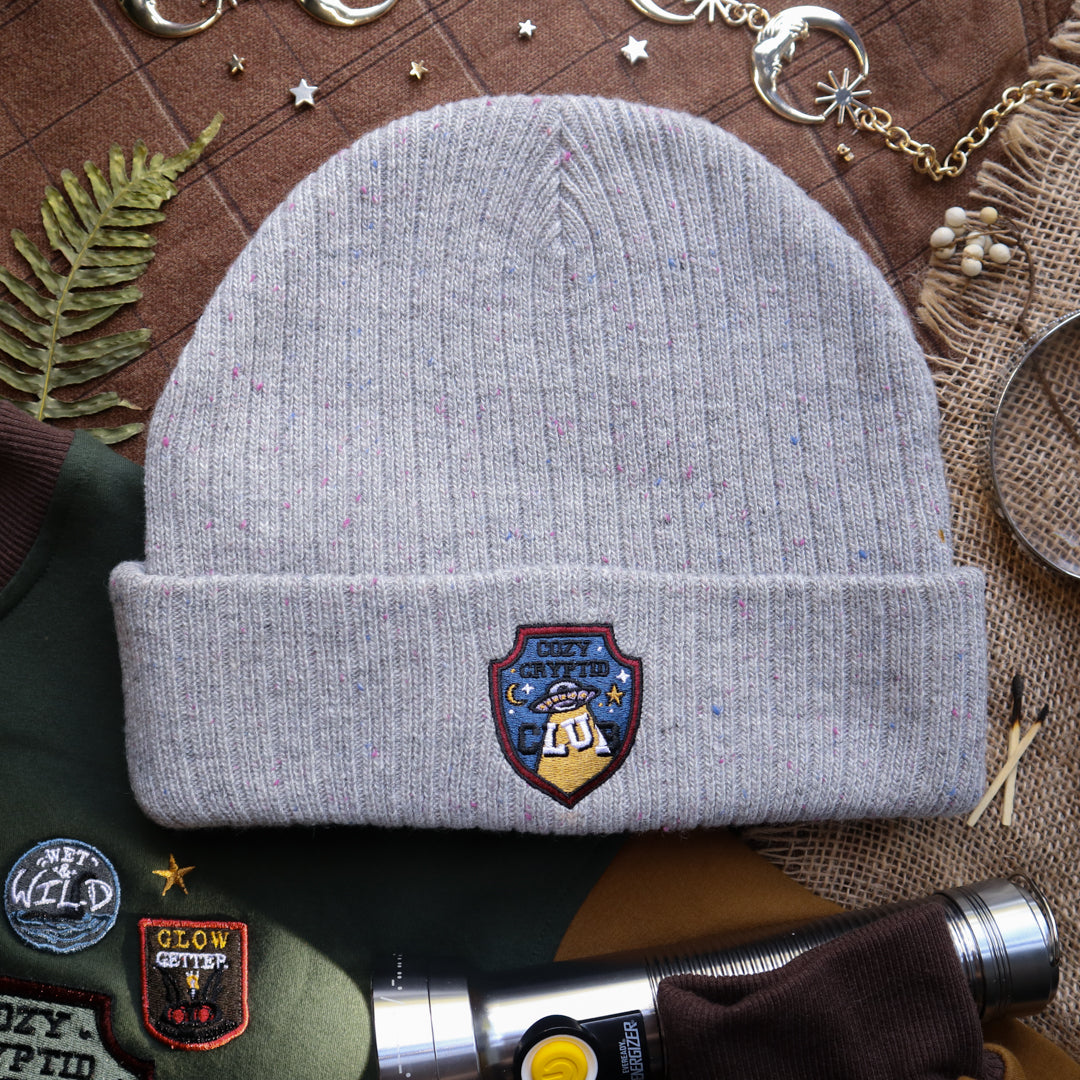 A cozy fog-grey cryptid camp-themed knit wool beanie with colourful speckles, embroidered with a cryptid club “iron on” patch, and Nessie monster.