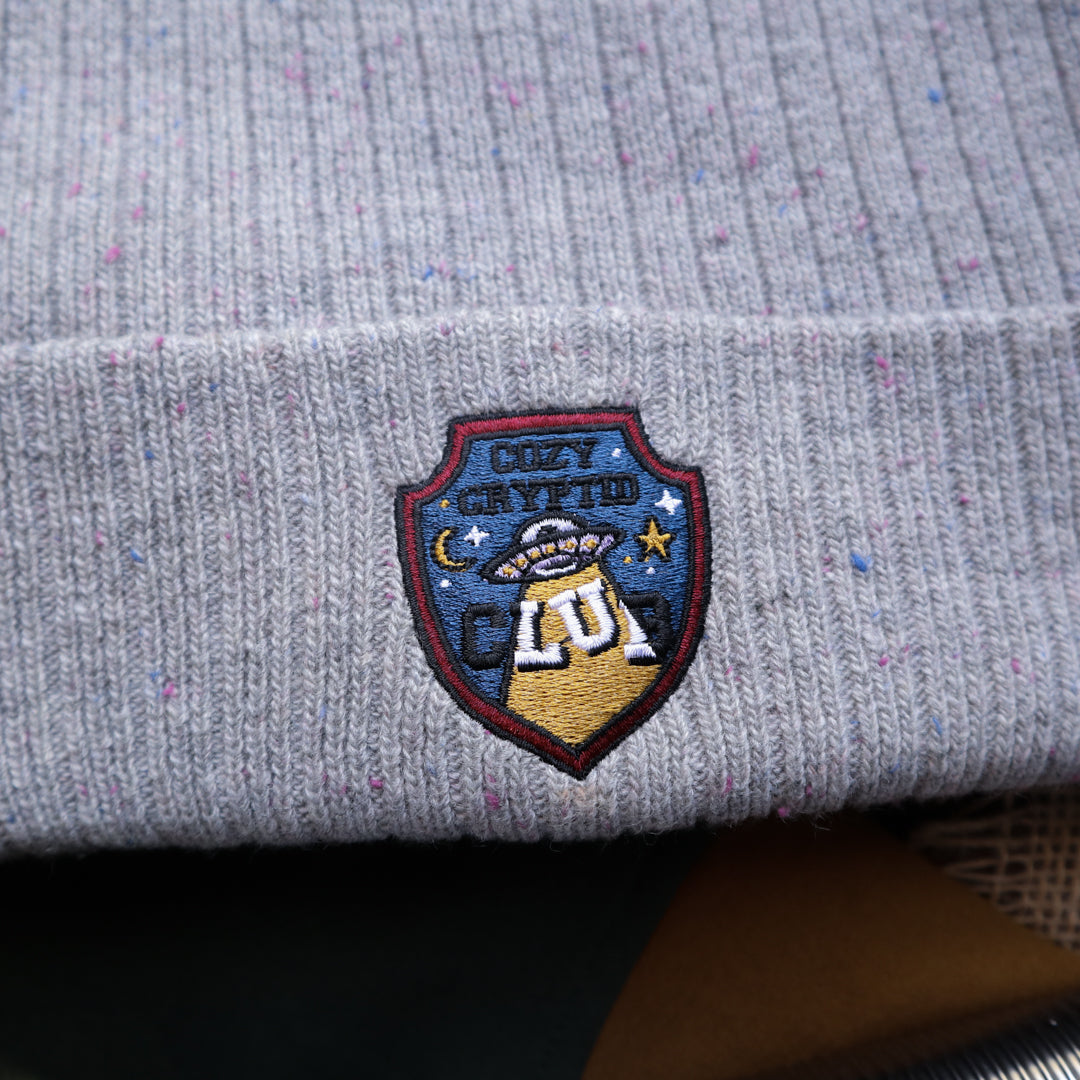 A cozy fog-grey cryptid camp-themed knit wool beanie with colourful speckles, embroidered with a cryptid club “iron on” patch, and Nessie monster.