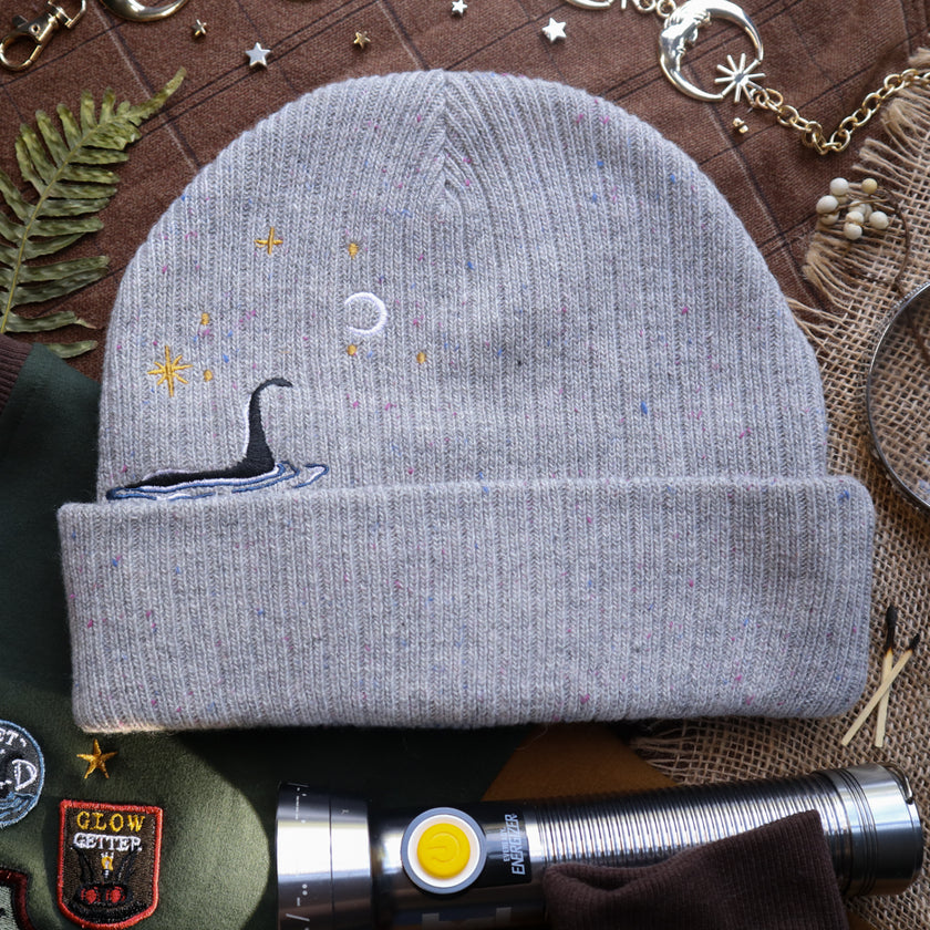 A cozy fog-grey cryptid camp-themed knit wool beanie with colourful speckles, embroidered with a cryptid club “iron on” patch, and Nessie monster.