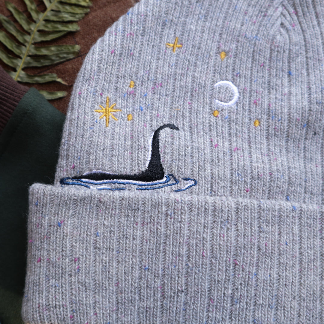 A cozy fog-grey cryptid camp-themed knit wool beanie with colourful speckles, embroidered with a cryptid club “iron on” patch, and Nessie monster.
