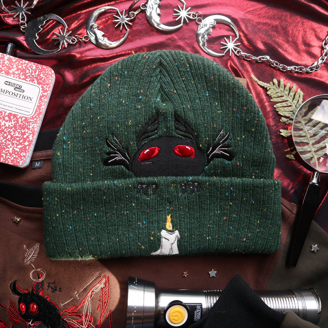 A cozy goblin-green knit wool beanie with colourful speckles, embroidered with a red-eyed, black-winged, flame-loving moth, and a glowing candle.