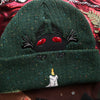 A cozy goblin-green knit wool beanie with colourful speckles, embroidered with a red-eyed, black-winged, flame-loving moth, and a glowing candle.