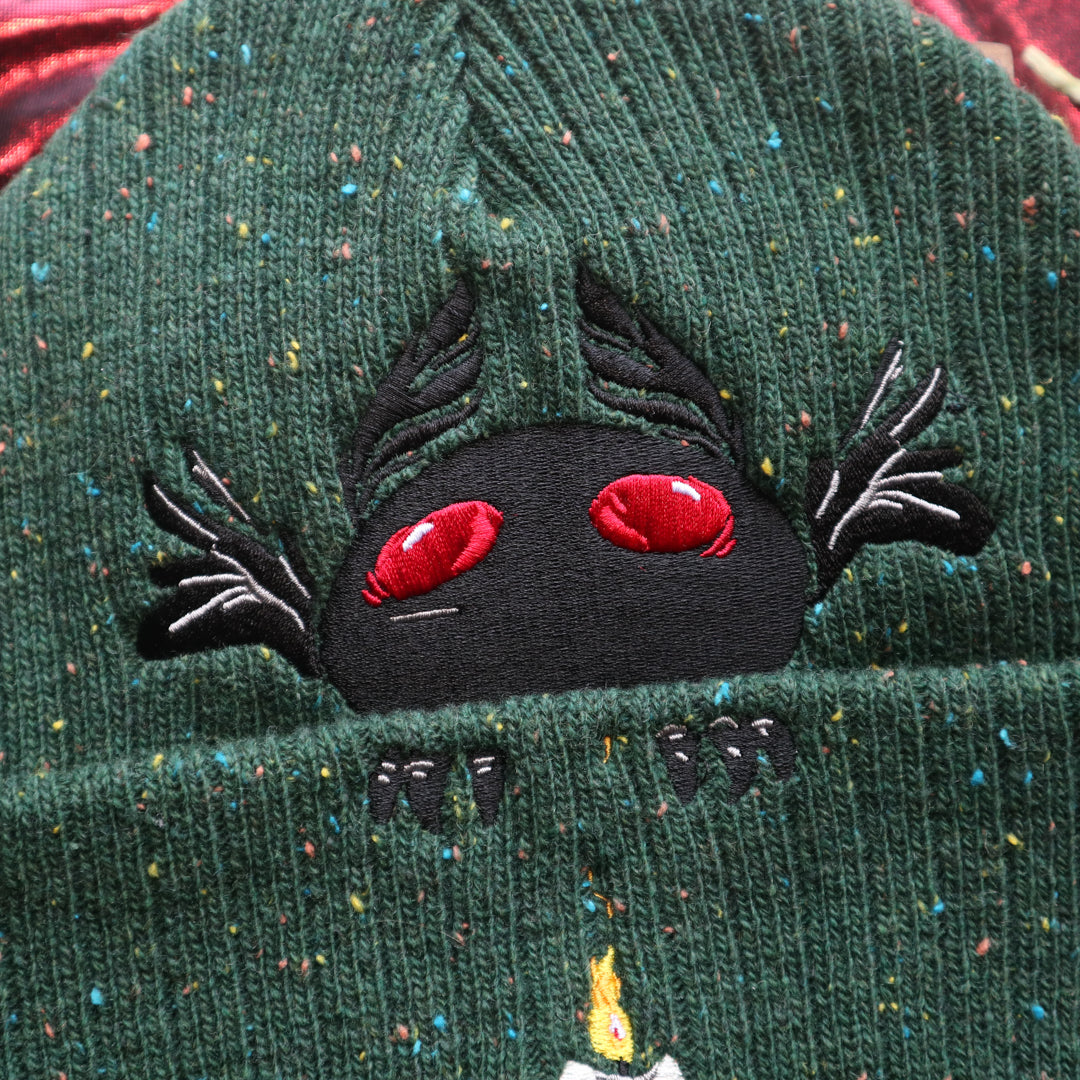 A cozy goblin-green knit wool beanie with colourful speckles, embroidered with a red-eyed, black-winged, flame-loving moth, and a glowing candle.