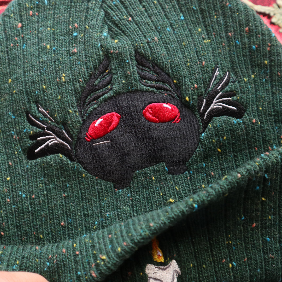 A cozy goblin-green knit wool beanie with colourful speckles, embroidered with a red-eyed, black-winged, flame-loving moth, and a glowing candle.