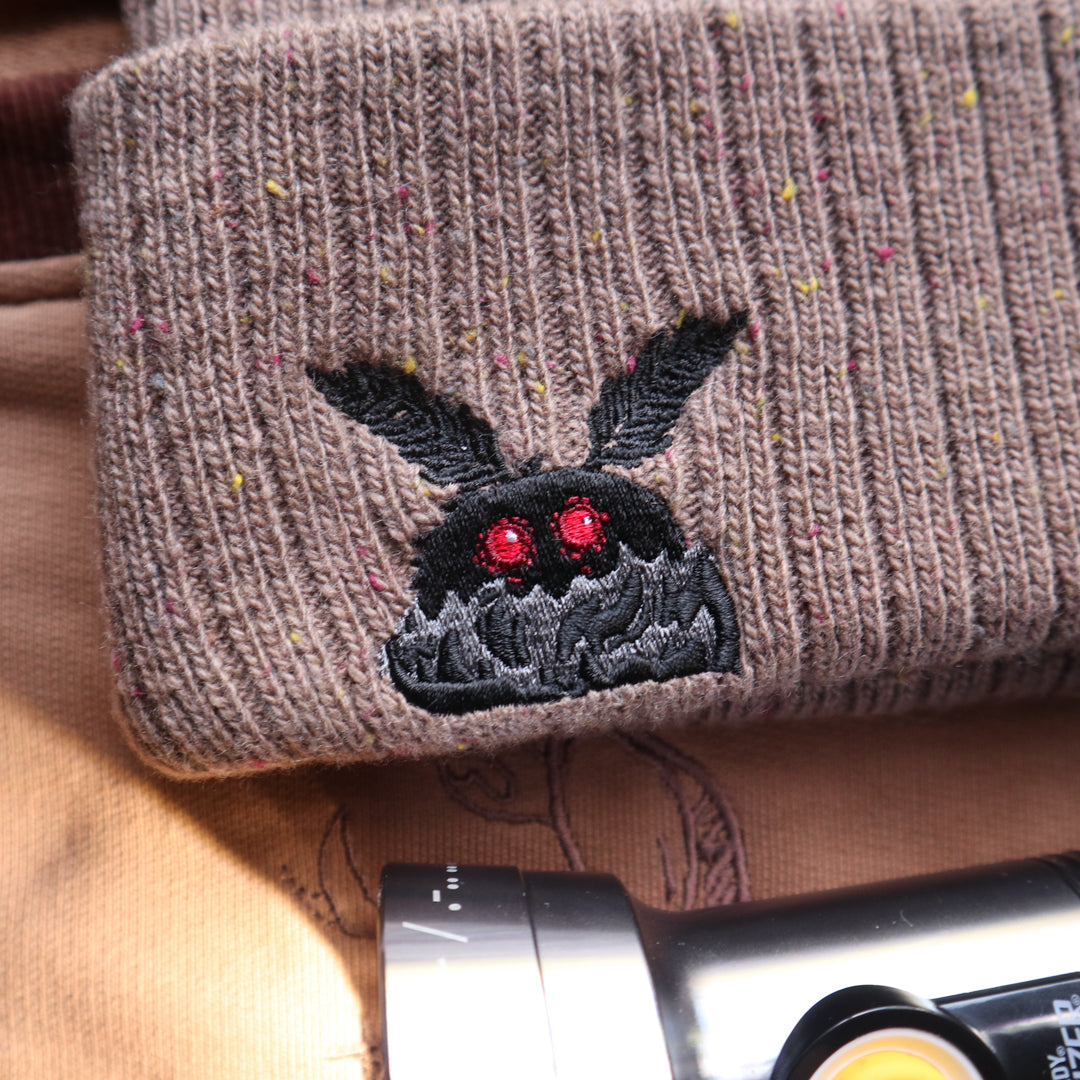 A cozy bone-dust brown cryptid-themed knit wool beanie with colourful speckles, embroidered with a tiny red-eyed, black-winged, flame-loving moth, and a secret message.