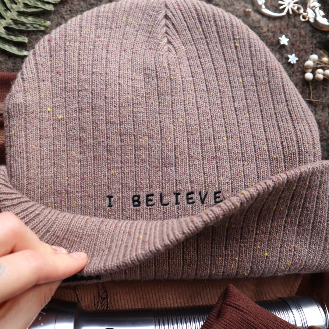 A cozy bone-dust brown cryptid-themed knit wool beanie with colourful speckles, embroidered with a tiny red-eyed, black-winged, flame-loving moth, and a secret message.