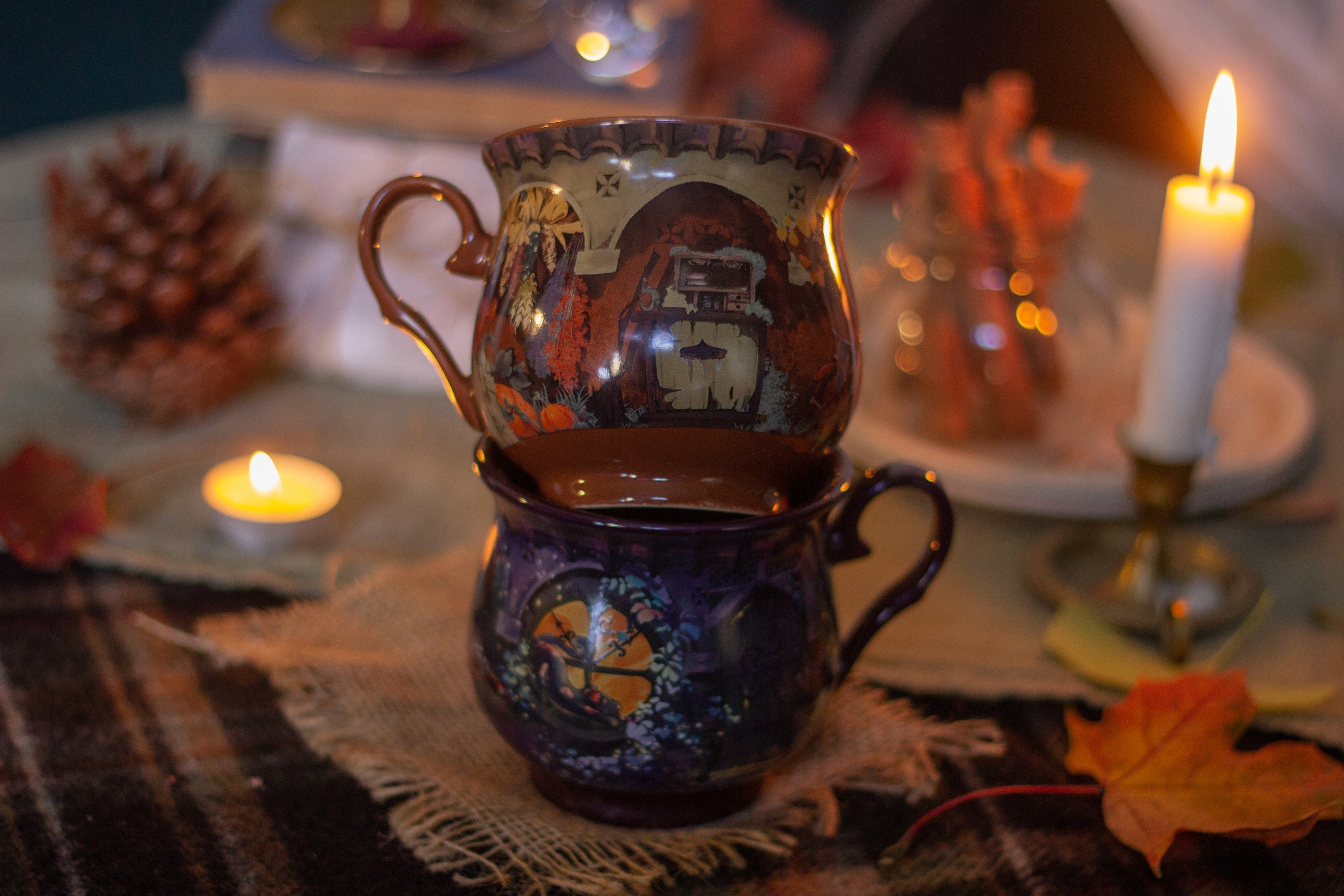 Two intricately designed witches cottage ceramic mugs, featuring vibrant seasonal and whimsical artwork. The top mug showcases an autumnal theme with pumpkins and fall foliage, while the bottom mug highlights an autumnal evening scene with a full moon, a prowling cat, and dark, enchanting colors.