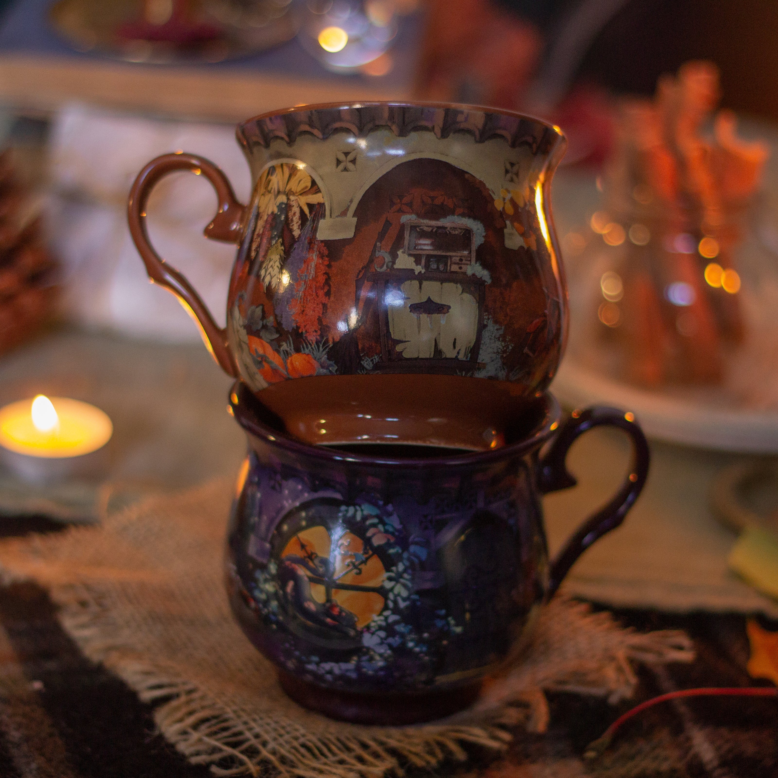 Two intricately designed witches cottage ceramic mugs, featuring vibrant seasonal and whimsical artwork. The top mug showcases an autumnal theme with pumpkins and fall foliage, while the bottom mug highlights an autumnal evening scene with a full moon, a prowling cat, and dark, enchanting colors.