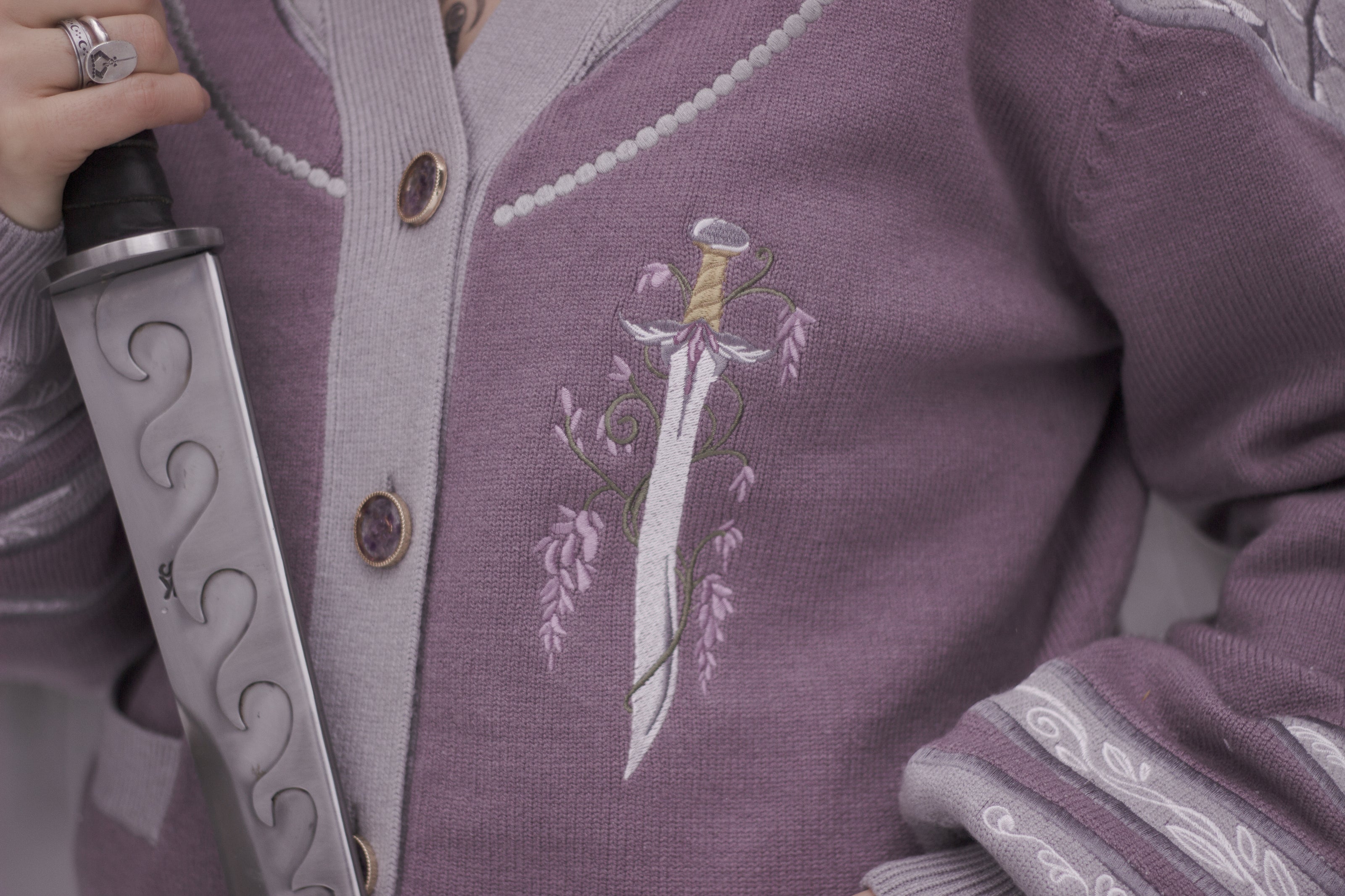 A soft button-up lavender and light plum knit cardigan shown in two styles: cropped and full-length. The design features intricate embroidery of swords and silver armour accents, evoking a medieval and fantasy-inspired look.