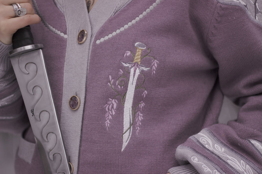 A soft button-up lavender and light plum knit cardigan shown in two styles: cropped and full-length. The design features intricate embroidery of swords and silver armour accents, evoking a medieval and fantasy-inspired look.