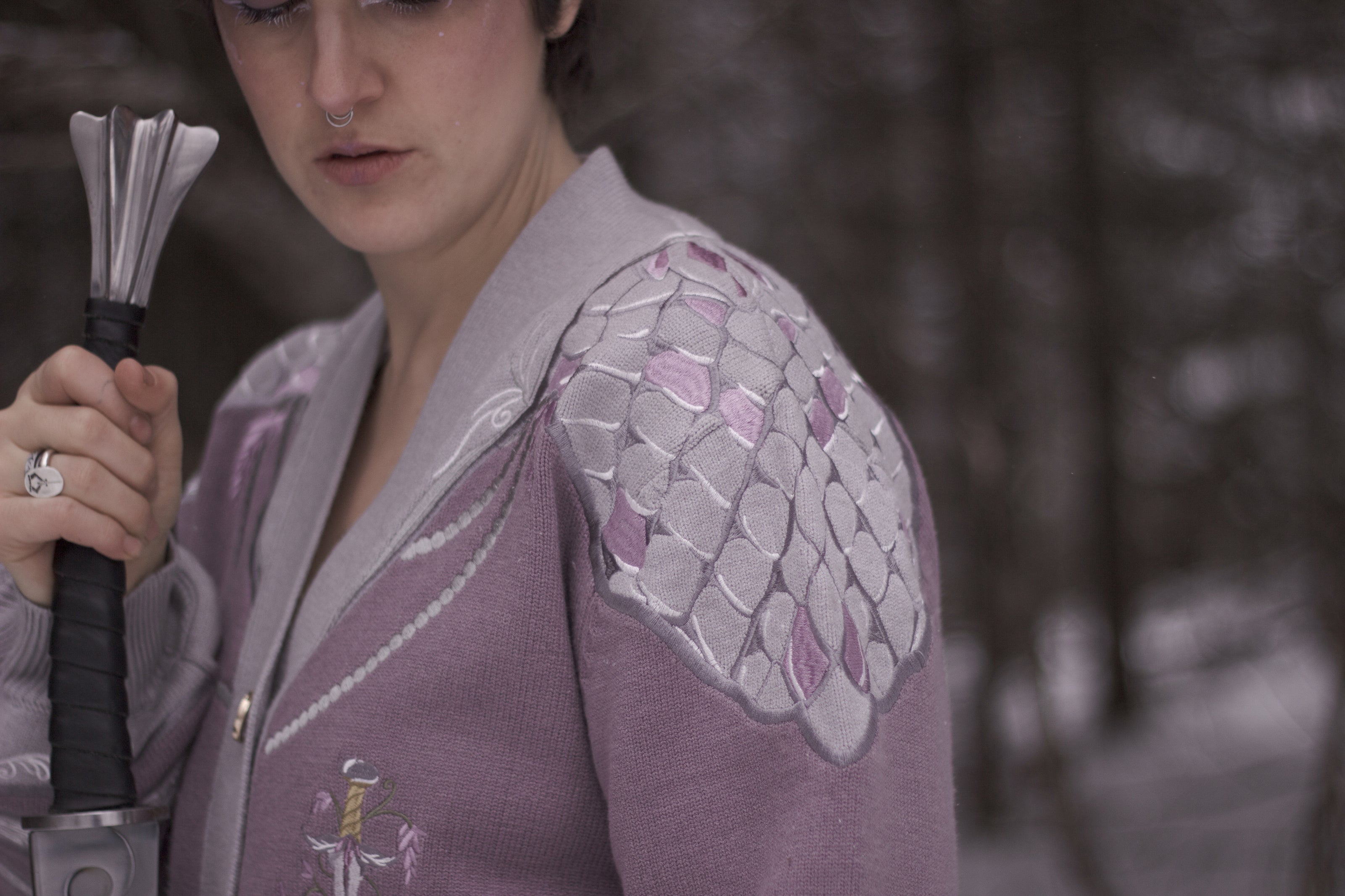 A soft button-up lavender and light plum knit cardigan shown in two styles: cropped and full-length. The design features intricate embroidery of swords and silver armour accents, evoking a medieval and fantasy-inspired look.