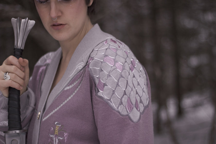 A soft button-up lavender and light plum knit cardigan shown in two styles: cropped and full-length. The design features intricate embroidery of swords and silver armour accents, evoking a medieval and fantasy-inspired look.