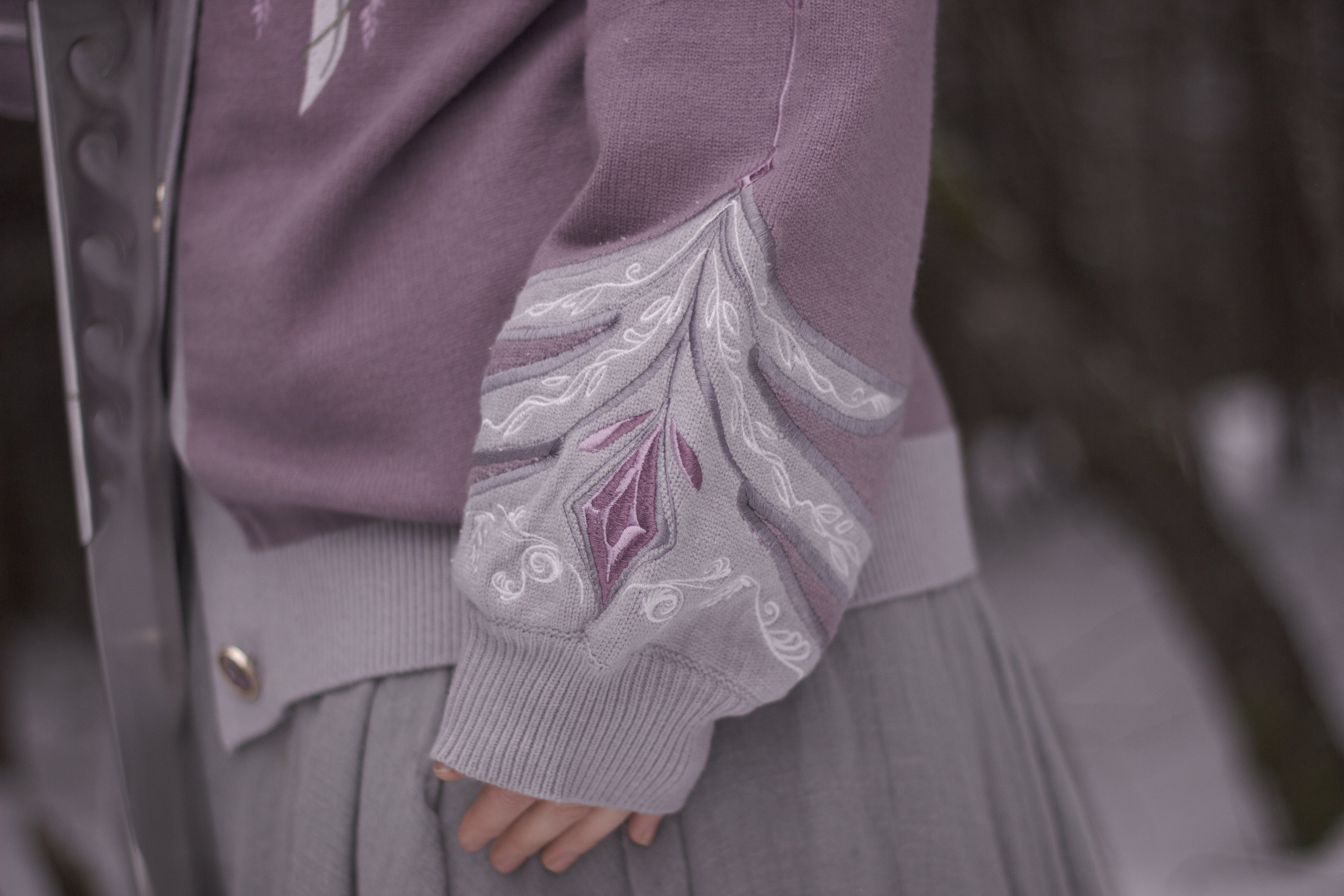 A soft button-up lavender and light plum knit cardigan shown in two styles: cropped and full-length. The design features intricate embroidery of swords and silver armour accents, evoking a medieval and fantasy-inspired look.
