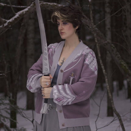 A soft button-up lavender and light plum knit cardigan shown in two styles: cropped and full-length. The design features intricate embroidery of swords and silver armour accents, evoking a medieval and fantasy-inspired look.